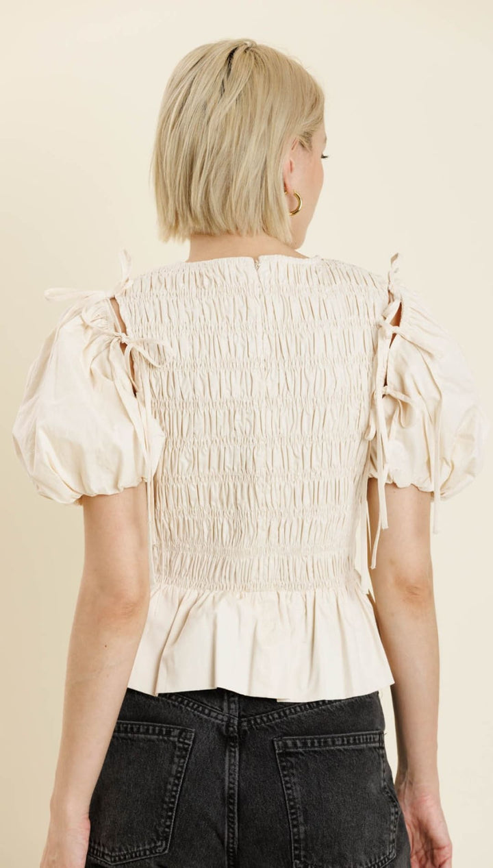 A woman’s off-white smocked peplum top with puff sleeves and tie details on the shoulders.