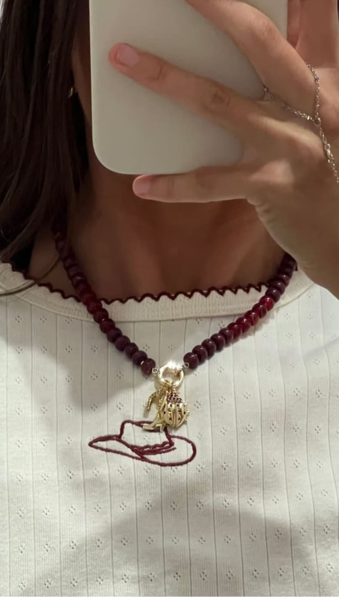 A woman wearing a red beaded necklace with a gold charm and a red embroidered cowboy hat on her white shirt, with a hand holding a phone to her face, wearing a delicate gold hand chain.
