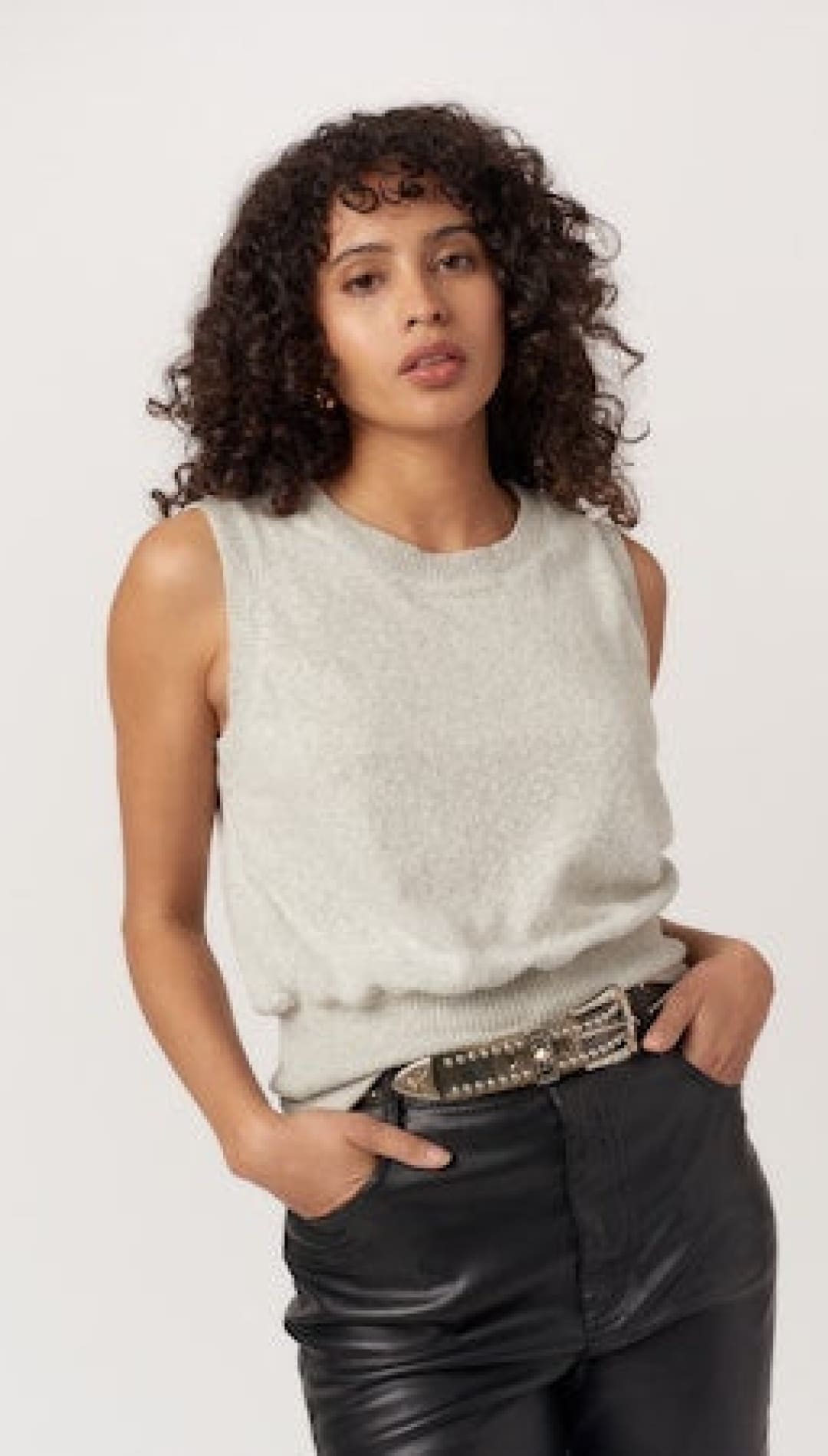 A sleeveless, light-colored knit top with a rounded neckline and a tucked-in waistline, paired with black leather-look pants and a studded belt.