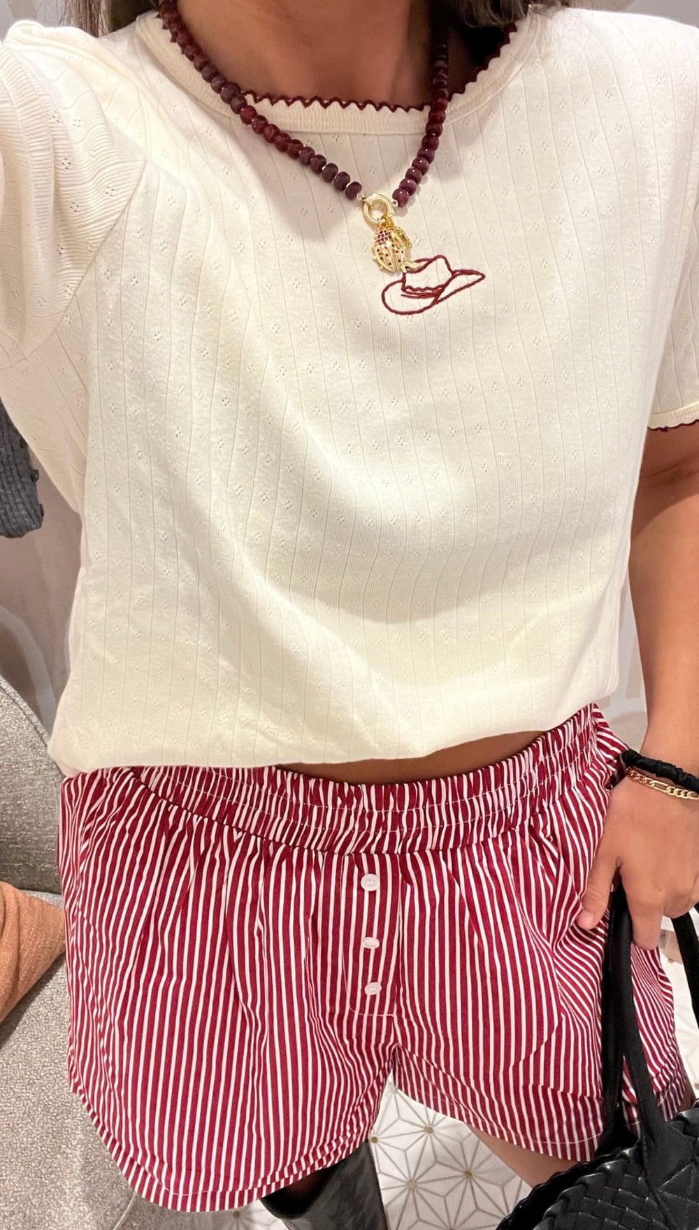A person wearing a cream-colored short-sleeved top with a red cowboy hat embroidery and red striped shorts.