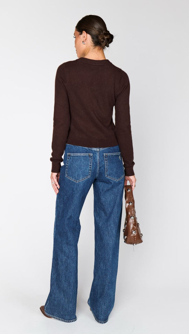A person wearing a long-sleeved dark brown top and dark blue wide-leg jeans, holding a brown studded handbag.