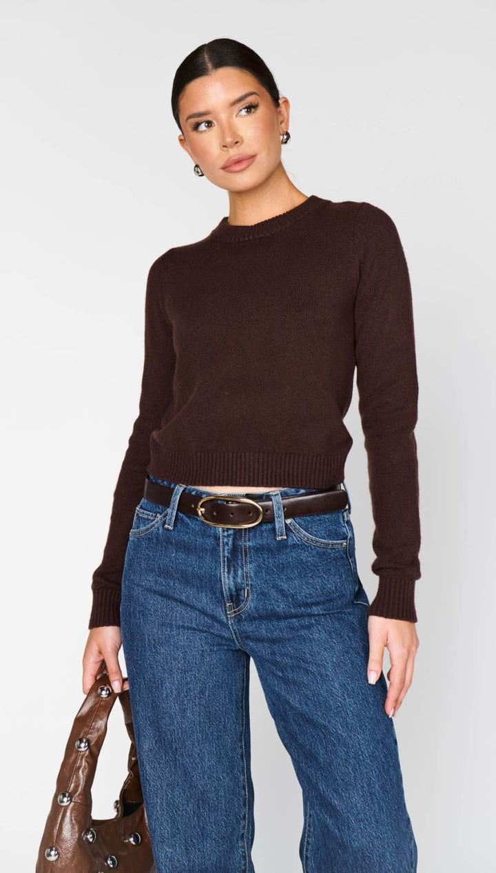 A woman wearing a dark brown long-sleeved crew-neck sweater, blue jeans, and a brown belt with a gold buckle, holding a brown studded handbag.