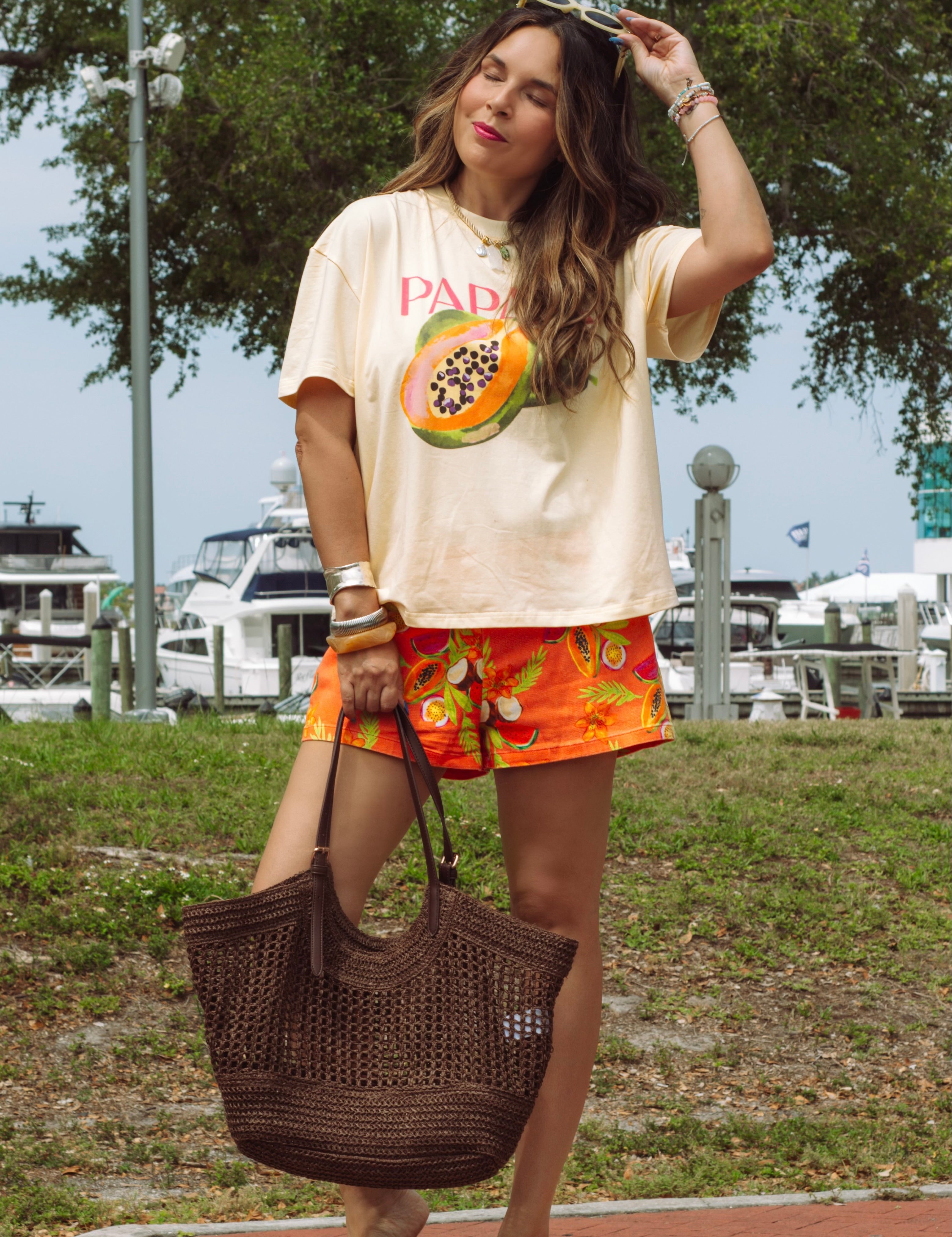Woman standing outdoors wearing a graphic t-shirt and colorful shorts, holding a brown bag.