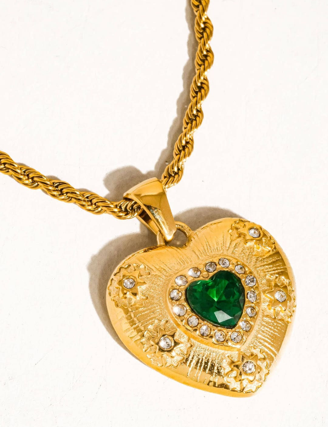 Gold heart-shaped pendant necklace with a central green gemstone surrounded by clear crystals and intricate engraved detailing.