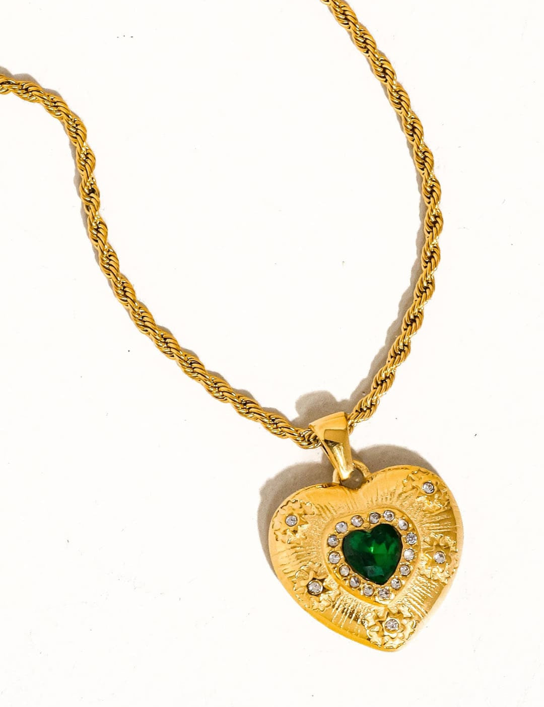 Gold heart-shaped pendant necklace with a central green gemstone surrounded by small clear stones and an ornate textured surface.