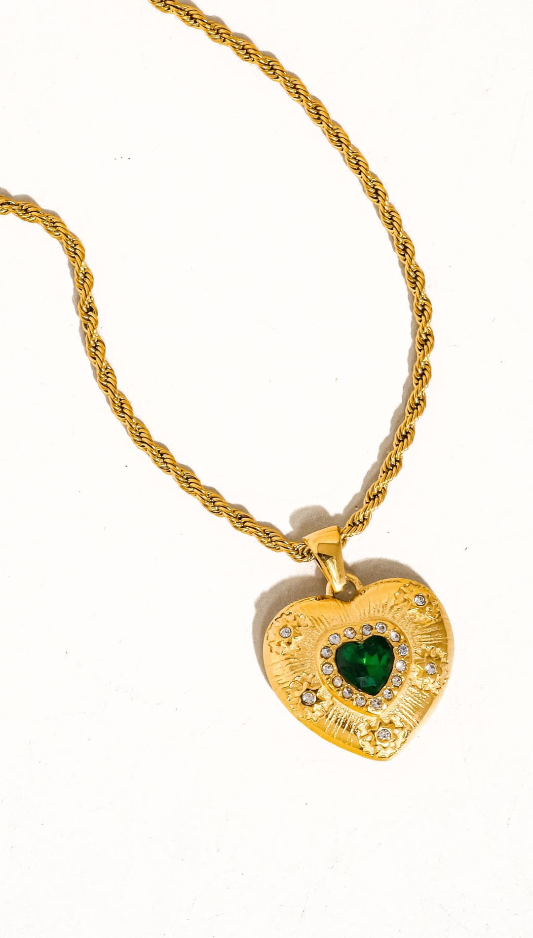 Gold heart-shaped pendant necklace with a central green gemstone surrounded by small clear stones and an ornate textured surface.
