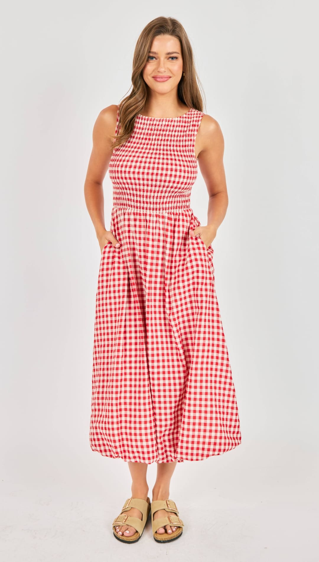 A red and white gingham sleeveless midi dress with a fitted bodice, smocked waistband, and two side pockets.
