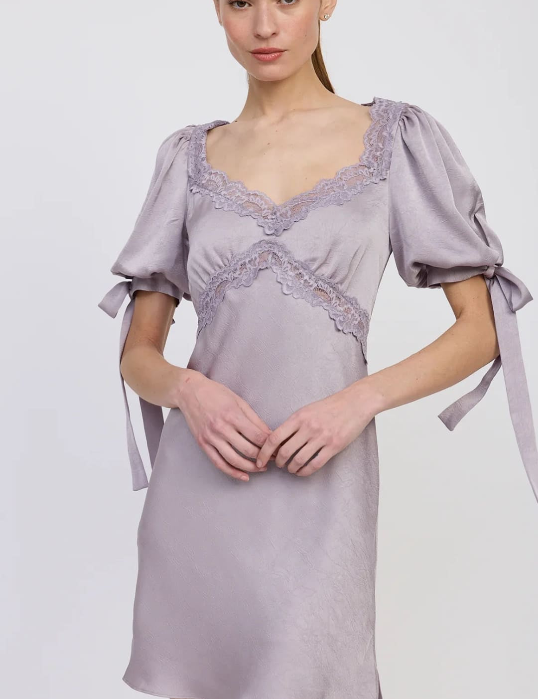 A woman modeling a short, light purple satin dress with puffy short sleeves, lace trim along the sweetheart neckline and bodice, and tie details on the sleeves.
