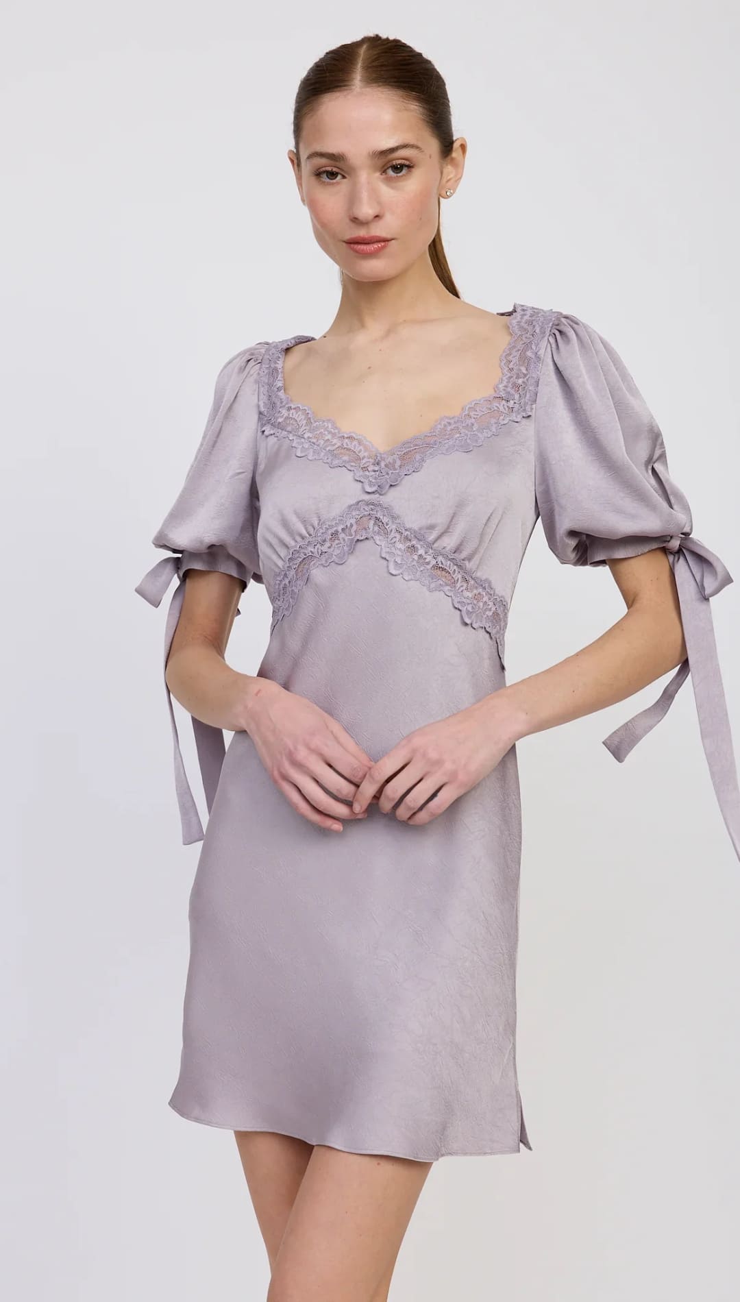 A woman modeling a short, light purple satin dress with puffy short sleeves, lace trim along the sweetheart neckline and bodice, and tie details on the sleeves.