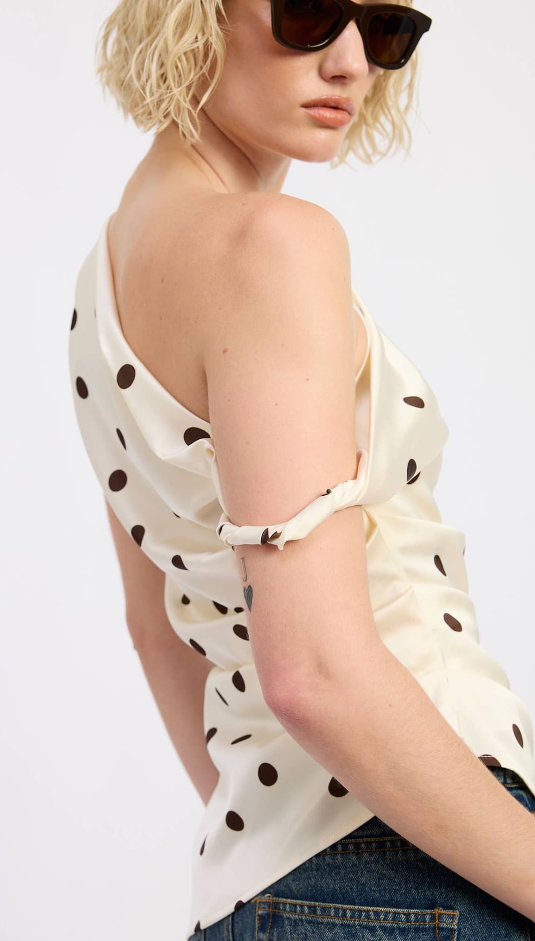 Off-white satin top with black polka dots and a one-shoulder design, featuring a tied knot at the arm.