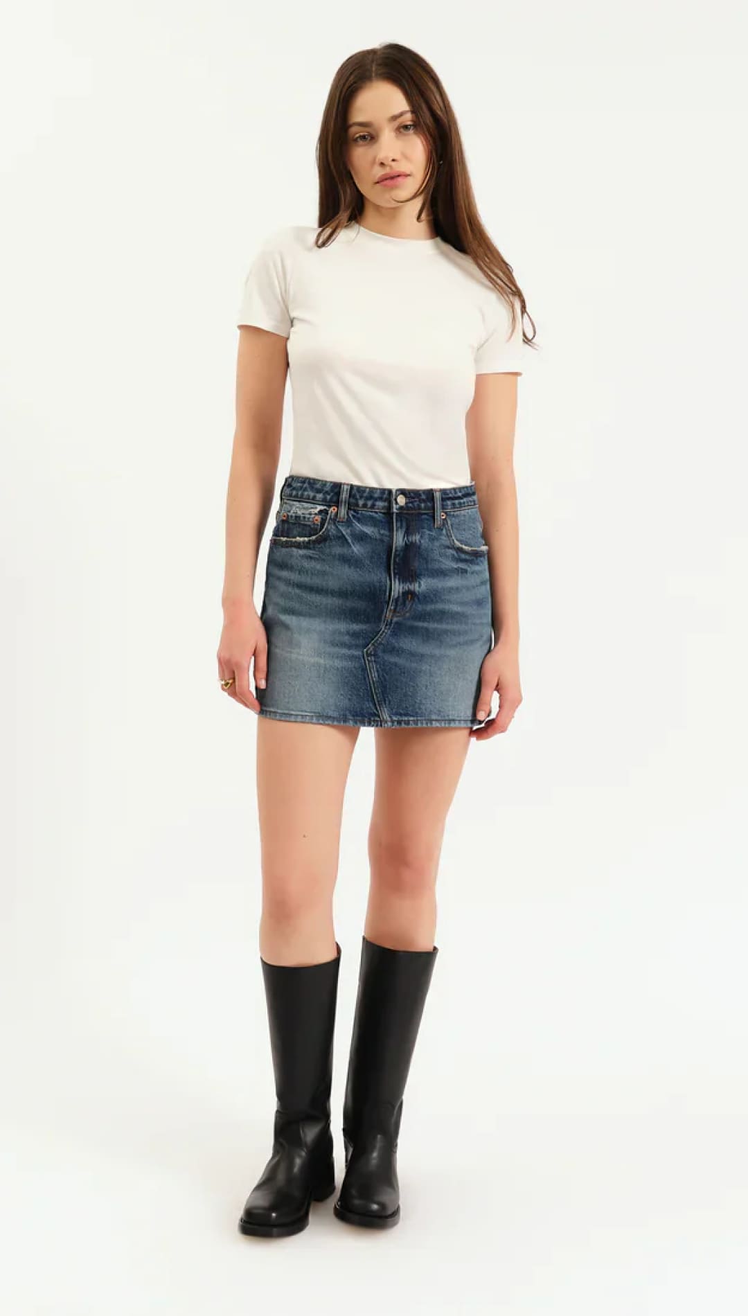 A woman wearing a white short-sleeve crewneck t-shirt, a blue denim mini skirt, and black knee-high boots.