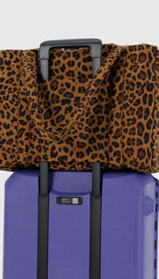 Leopard print travel tote bag with black handles and trim, placed on purple rolling suitcase.