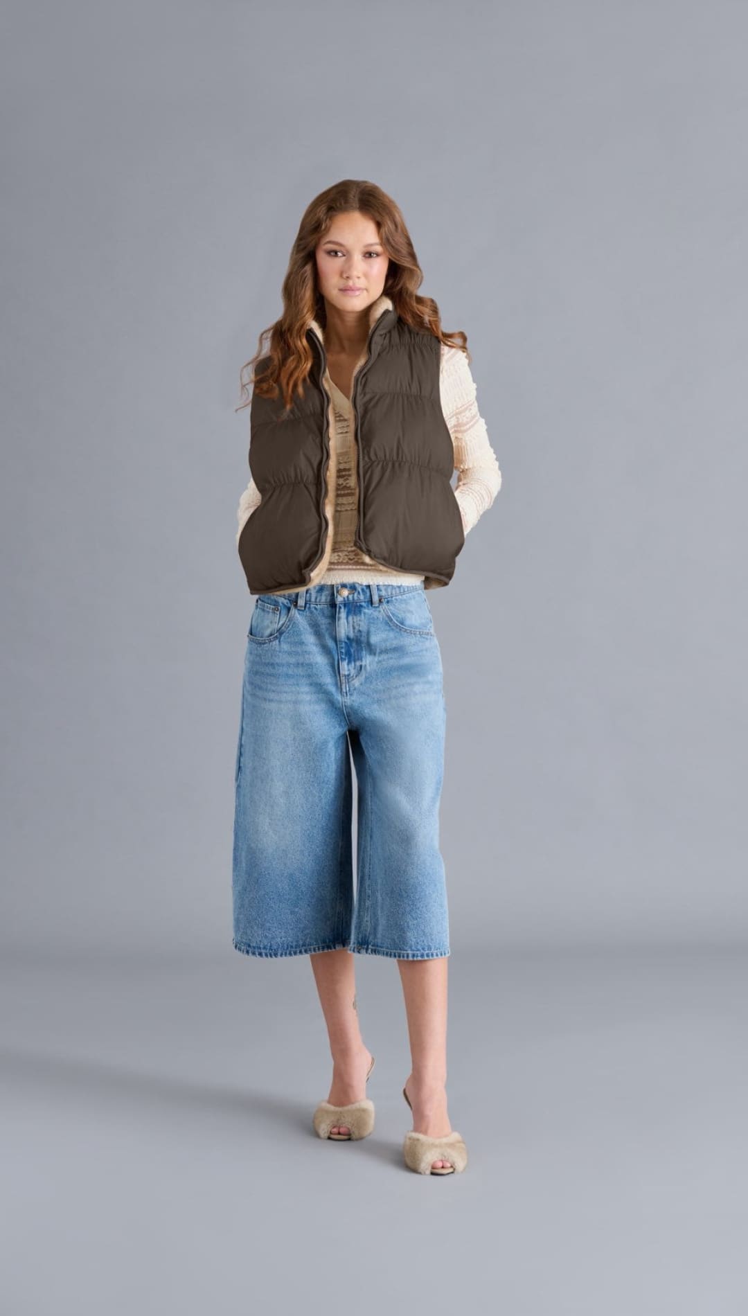 A young woman models a dark olive green puffer vest, light beige long-sleeved shirt, wide-leg denim culottes, and beige open-toed slip-on heels.