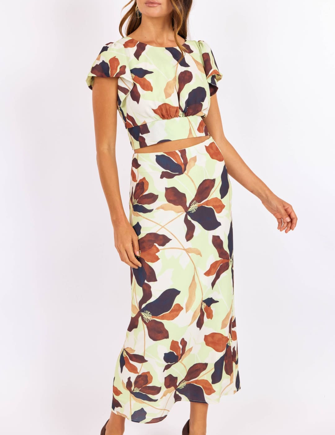 A woman wears a two-piece floral set featuring short flutter sleeves and a high-waisted midi skirt, with bold leaf patterns in earthy tones of brown, navy, and cream on a light mint background.