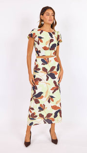 A woman wears a two-piece floral print outfit featuring short sleeves and a mid-thigh length skirt, with leaves in shades of brown, navy blue, and rust on an off-white background.