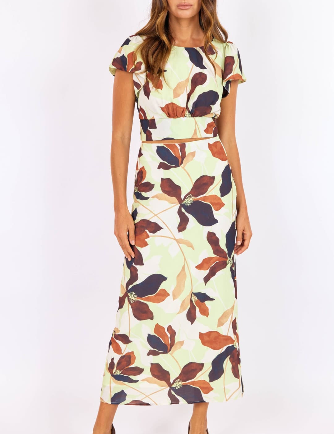 A woman wears a two-piece floral dress with short sleeves and a matching belt, featuring leaves in shades of brown, navy blue, and orange on an off-white background.