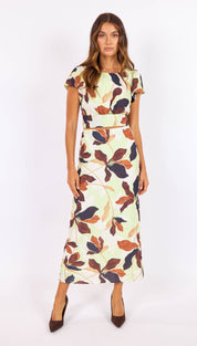 A woman wears a two-piece floral dress with short sleeves and a matching belt, featuring leaves in shades of brown, navy blue, and orange on an off-white background.