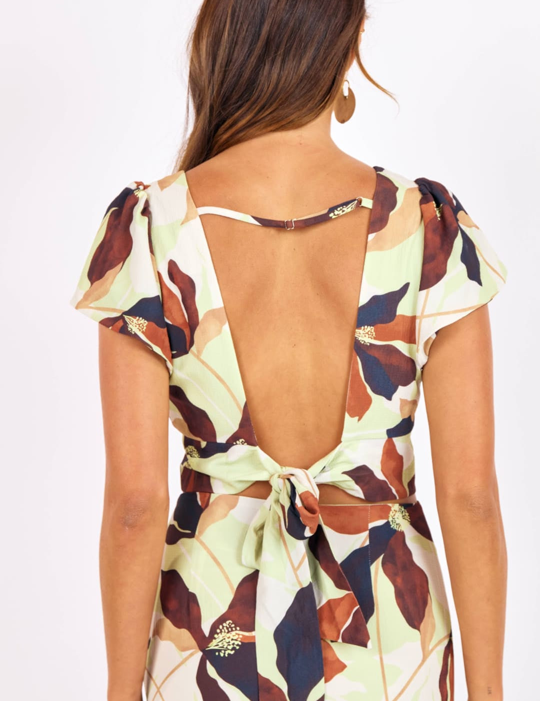 A woman wears a light green dress with a floral pattern in shades of brown, navy blue, and beige, featuring short puffed sleeves, an open back tied at the waist with fabric ties adorned with small metallic embellishments.