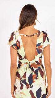 A woman wears a light green dress with a floral pattern in shades of brown, navy blue, and beige, featuring short puffed sleeves, an open back tied at the waist with fabric ties adorned with small metallic embellishments.