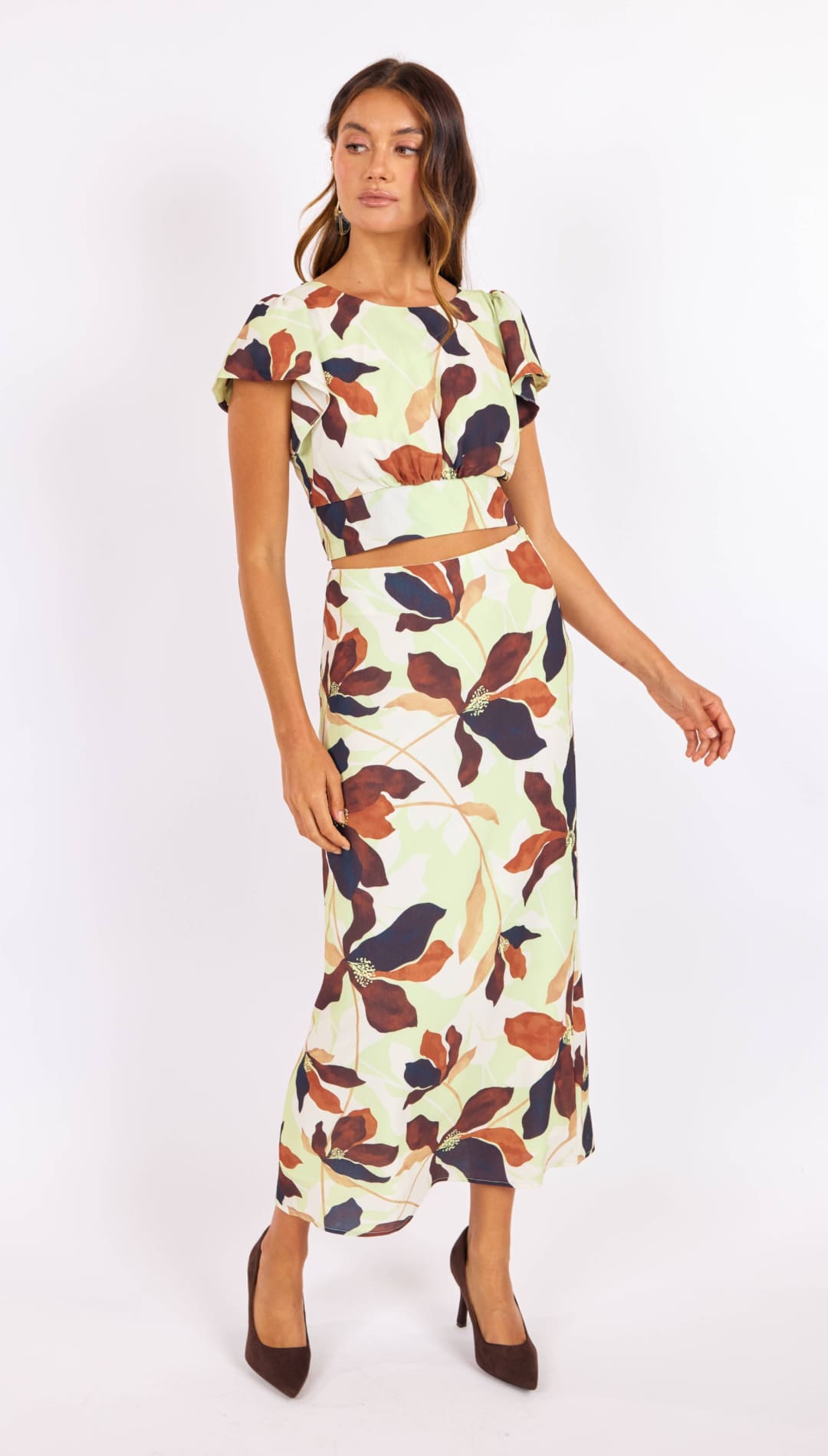 A woman wears a two-piece floral set featuring short flutter sleeves and a high-waisted midi skirt, with bold leaf patterns in earthy tones of brown, navy, and cream on a light mint background.