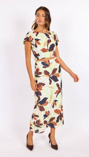 A woman wears a two-piece floral set featuring short flutter sleeves and a high-waisted midi skirt, with bold leaf patterns in earthy tones of brown, navy, and cream on a light mint background.