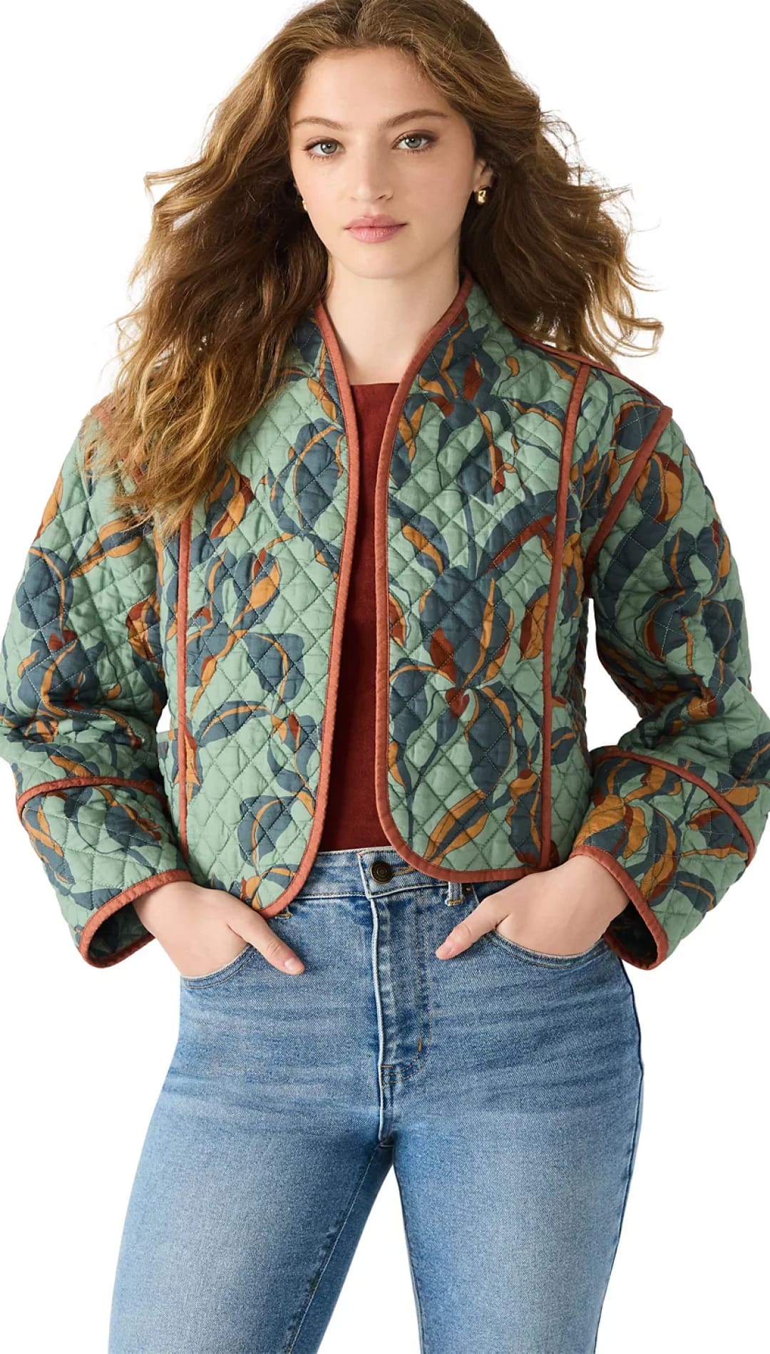 A woman modeling a quilted, cropped, floral-patterned jacket with rust-colored trim over a red top and light wash jeans.