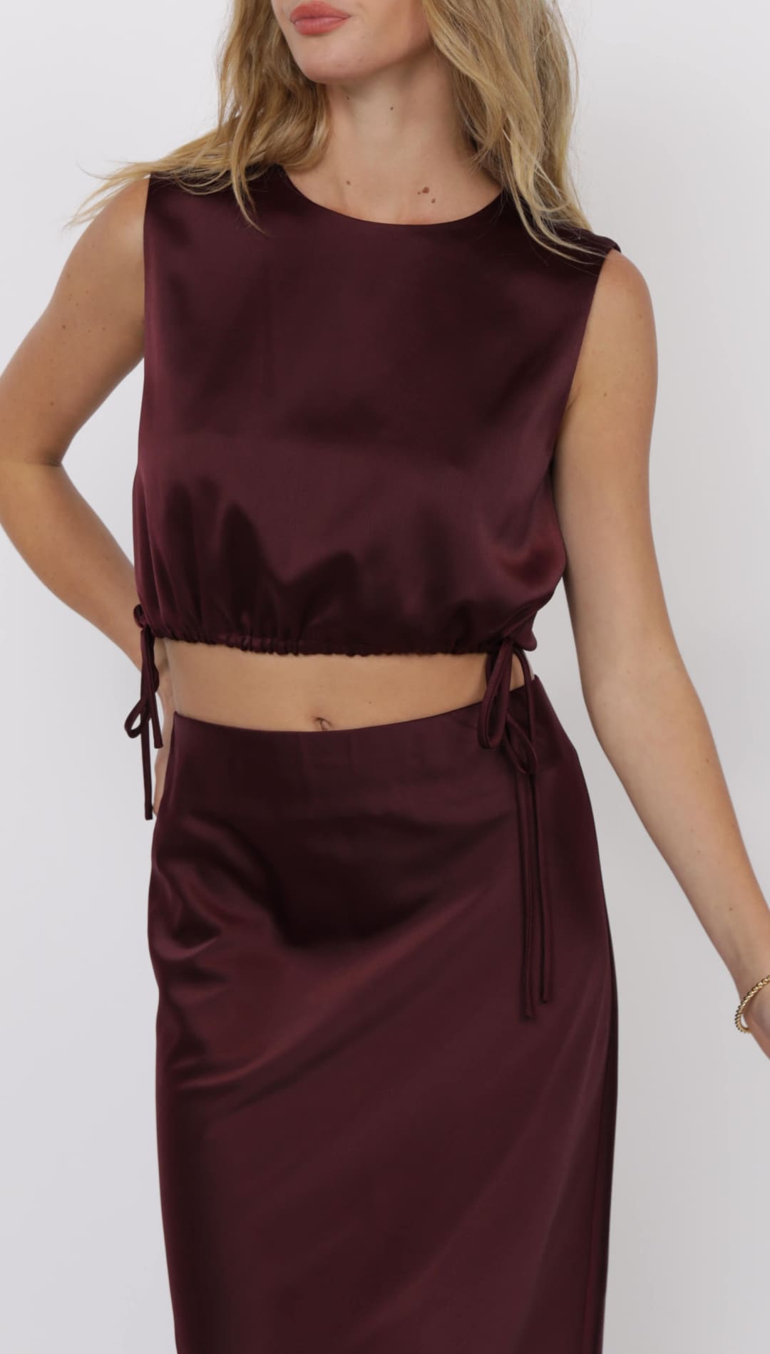 A two-piece satin co-ord set in a rich burgundy color, featuring a sleeveless crop top with adjustable drawstrings at the hem and a matching midi skirt.