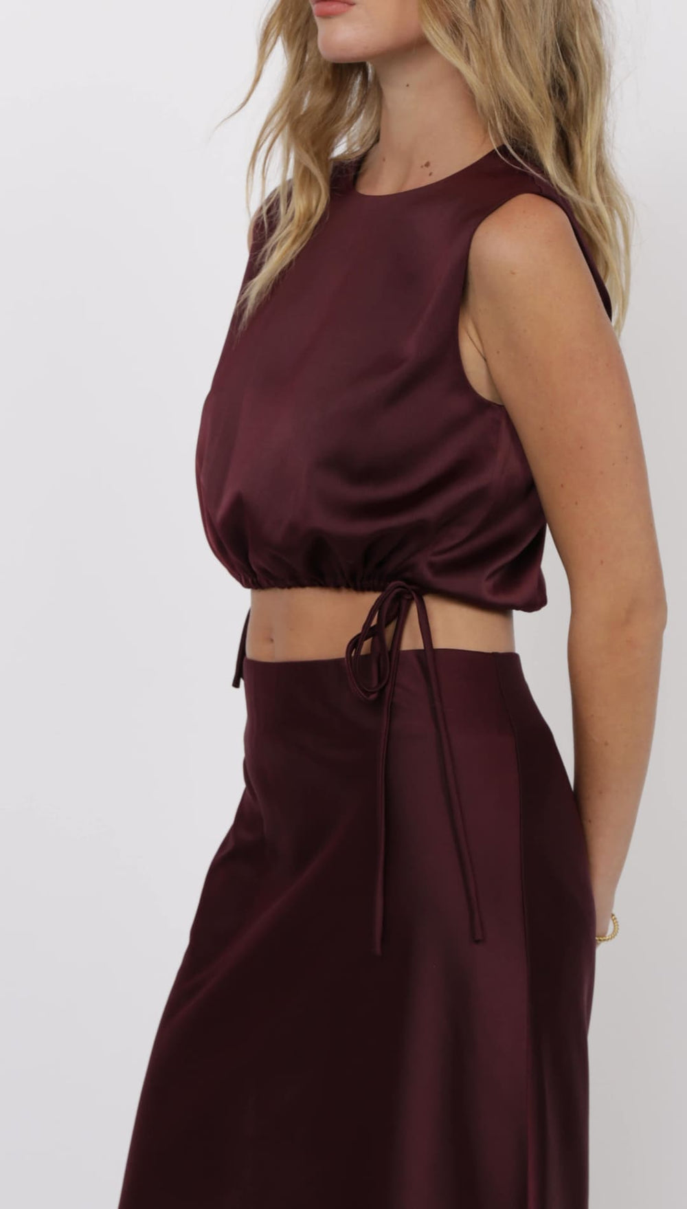 A women’s sleeveless, crew-neck, cropped satin top with a drawstring hem, paired with a matching satin midi skirt.