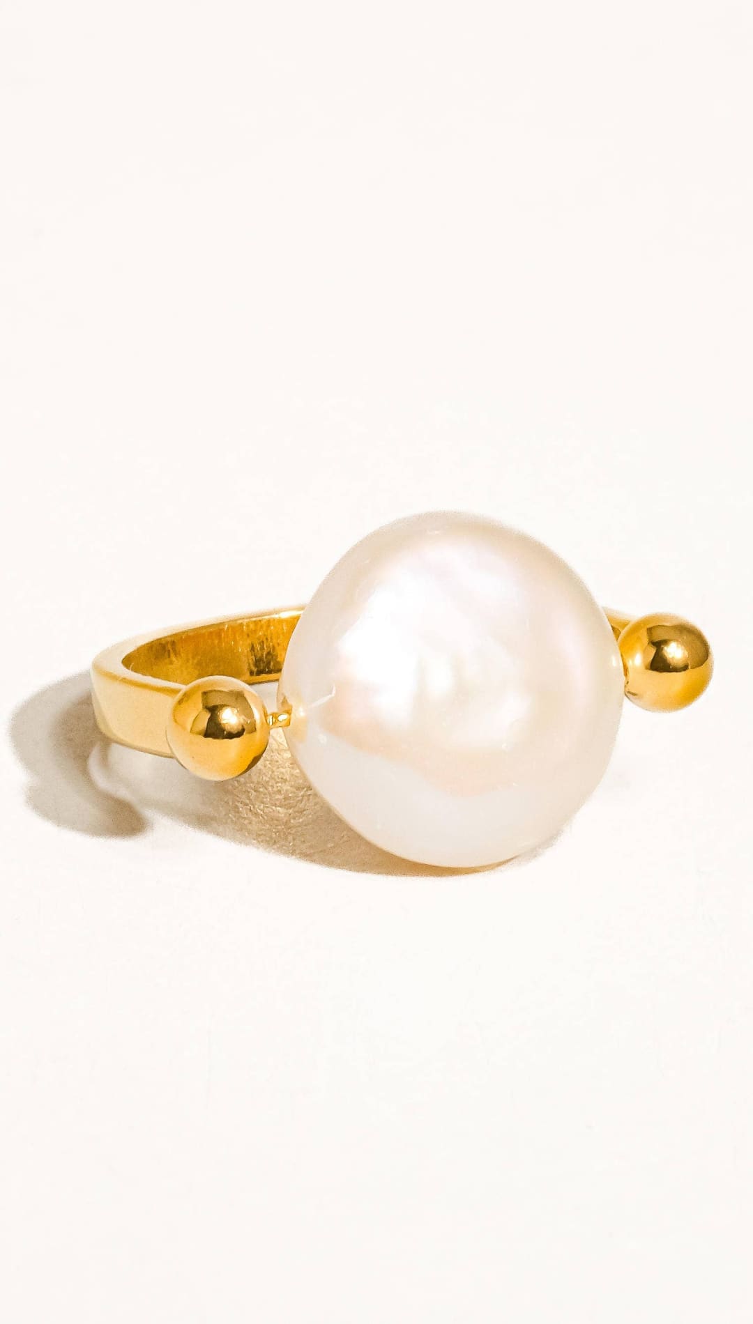 A gold-toned ring featuring a large, lustrous white pearl flanked by two small golden spheres.