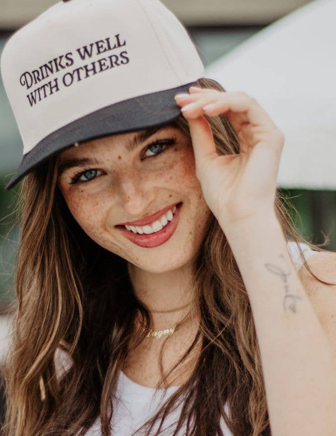 White and gray baseball cap with ’drinks well with others’ text.