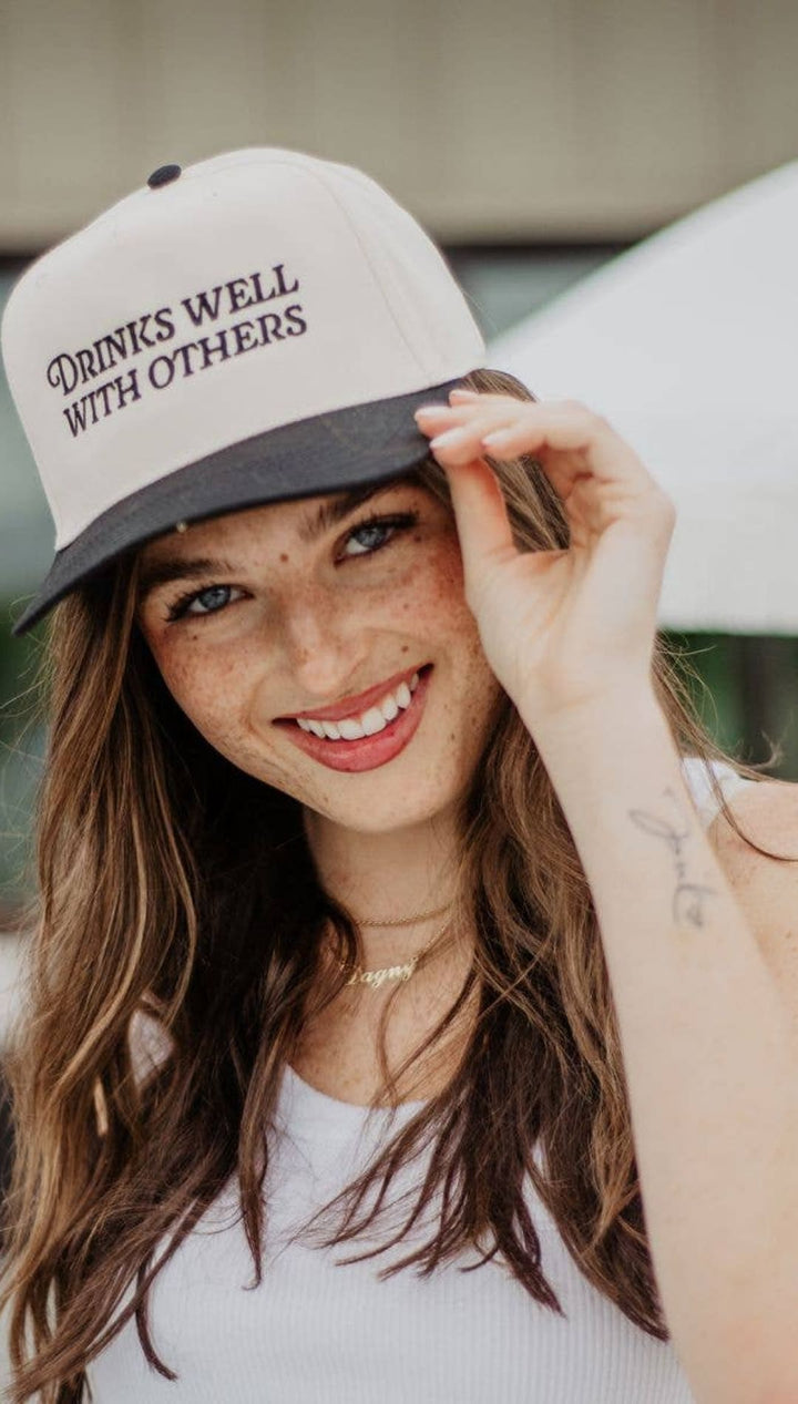 White and gray baseball cap with ’drinks well with others’ text.