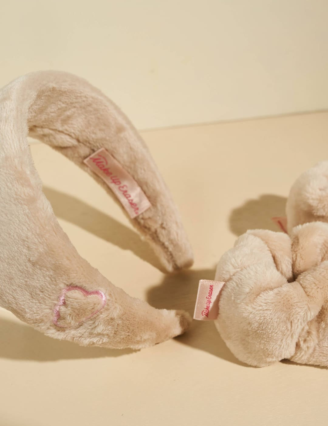 Cream-colored plush scrunchie with pink accents and bow design, likely from a popular fashion brand.
