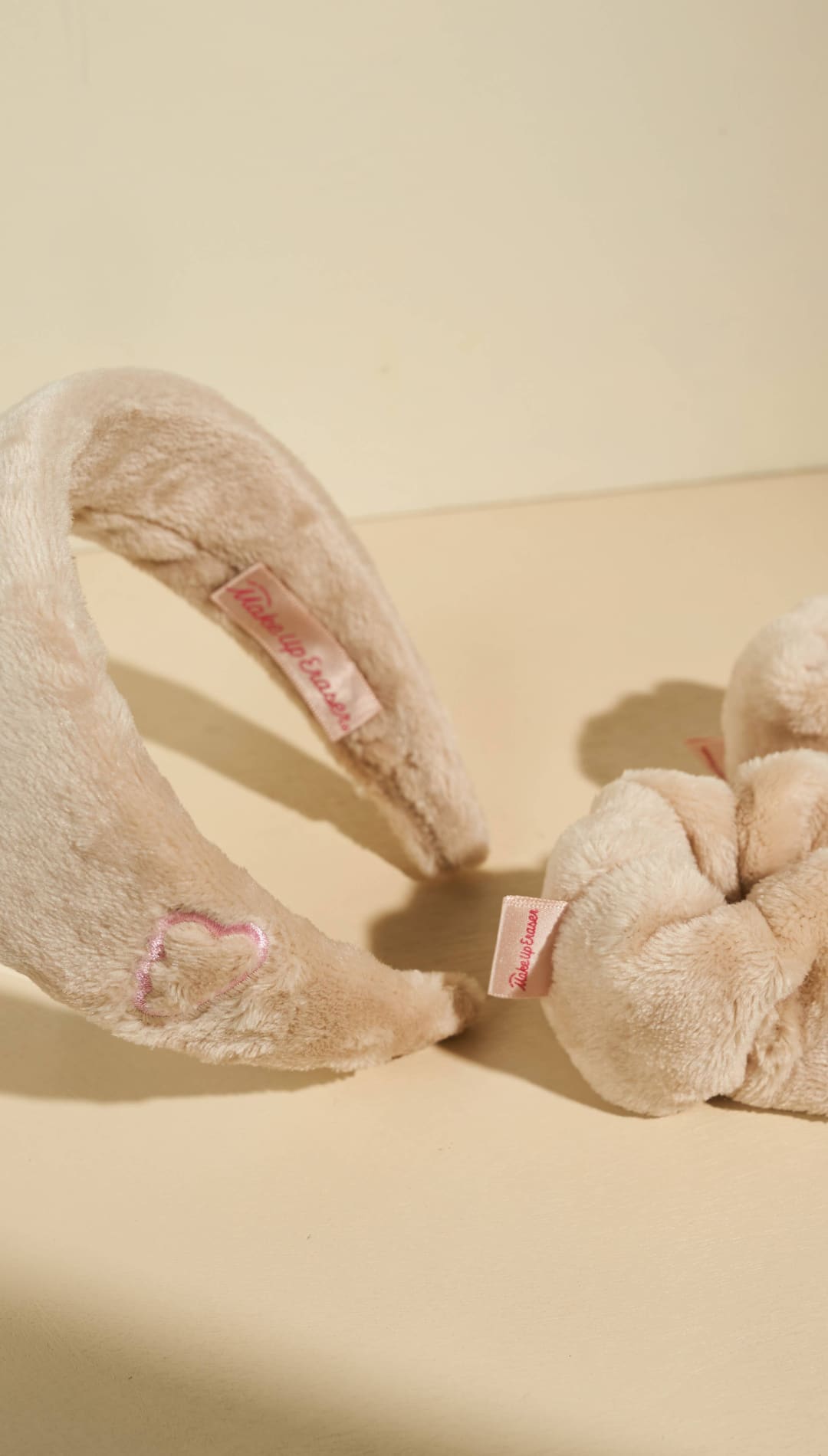 Cream-colored plush scrunchie with pink accents and bow design, likely from a popular fashion brand.