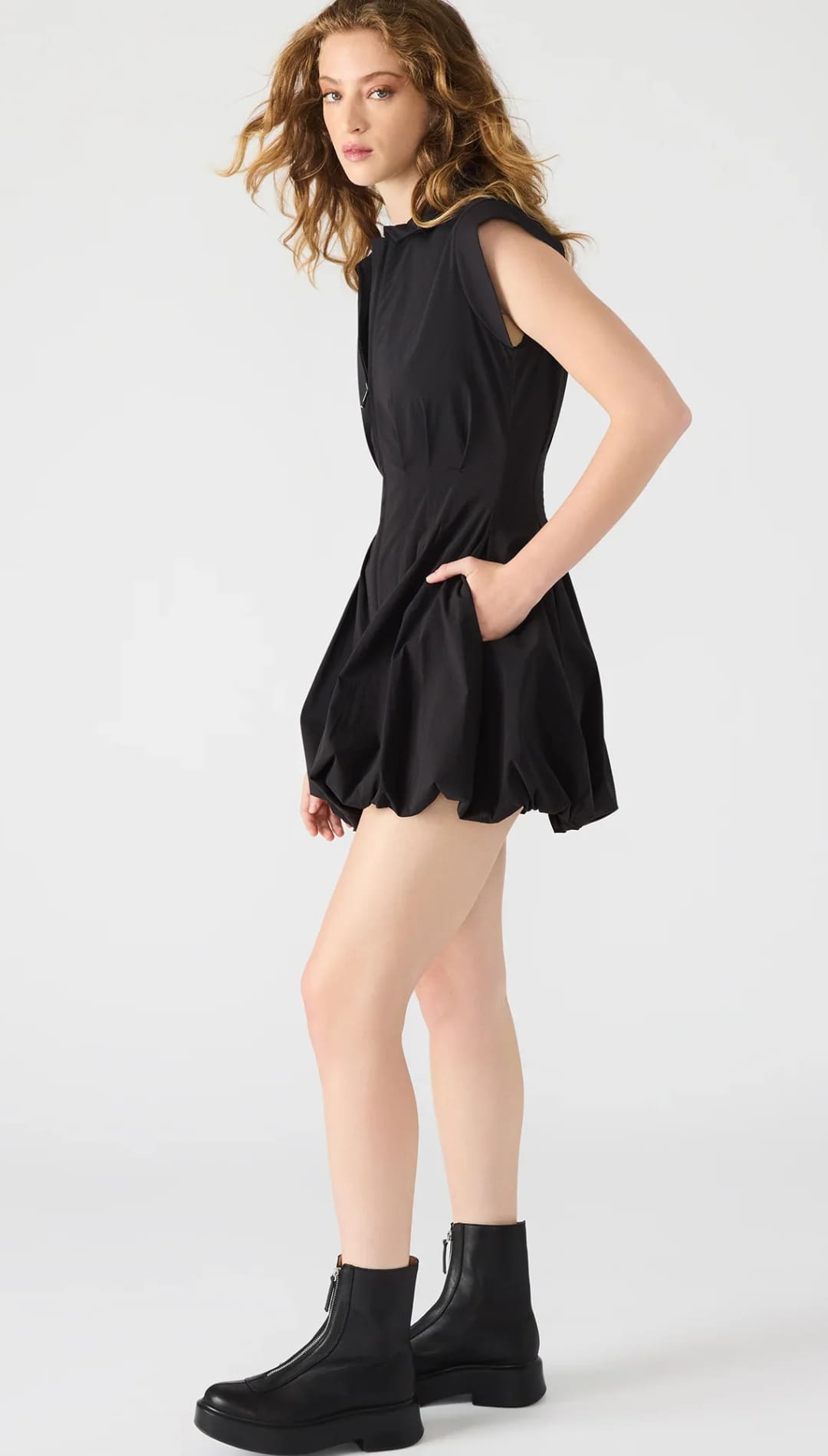 A black sleeveless mini dress with a fitted bodice, a gathered, voluminous skirt, and front button closure, paired with black combat boots.