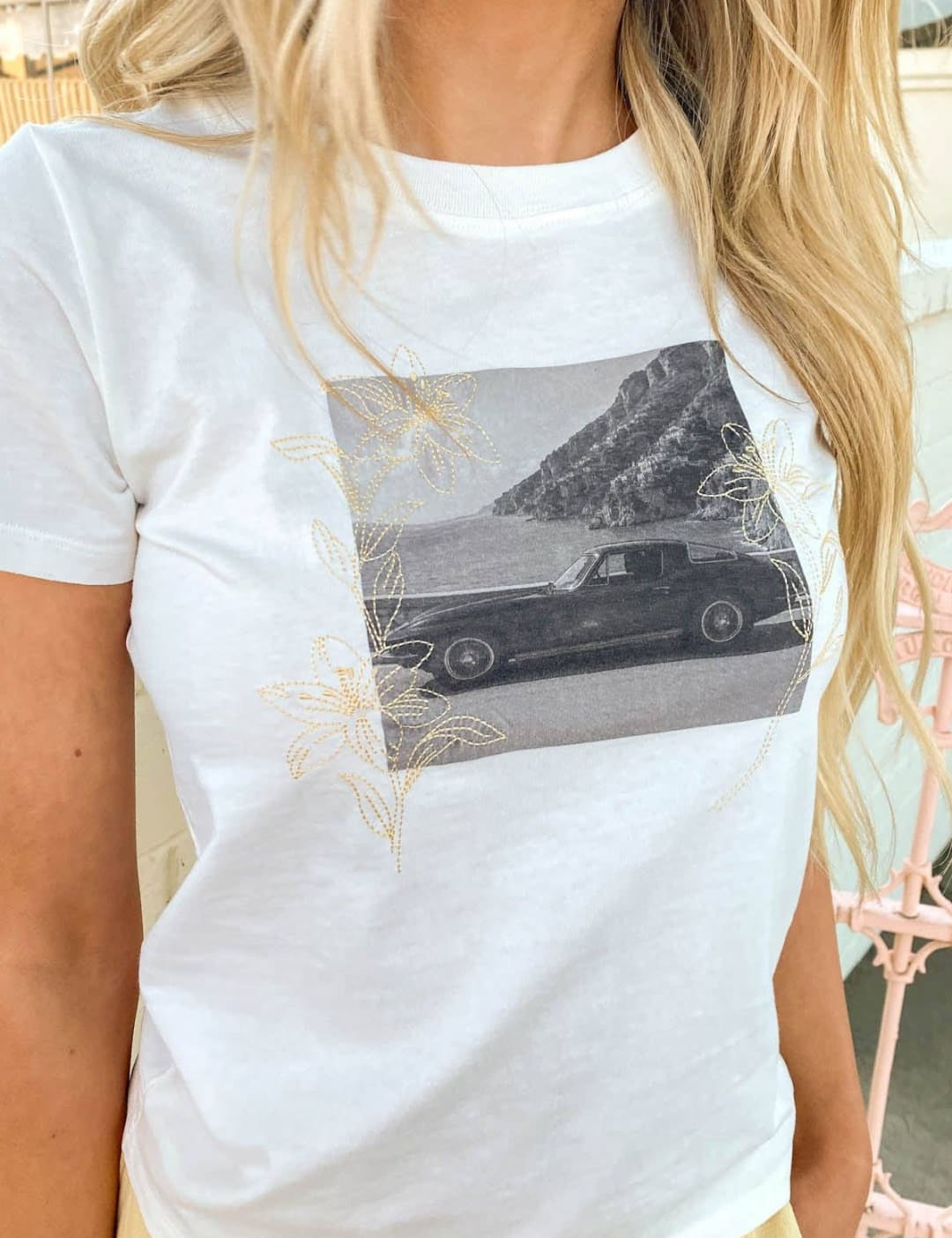 White short-sleeved t-shirt featuring a black-and-white graphic of a classic car on a coastal cliff, accented with delicate gold foil floral designs.