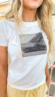 White short-sleeved t-shirt featuring a black-and-white graphic of a classic car on a coastal cliff, accented with delicate gold foil floral designs.