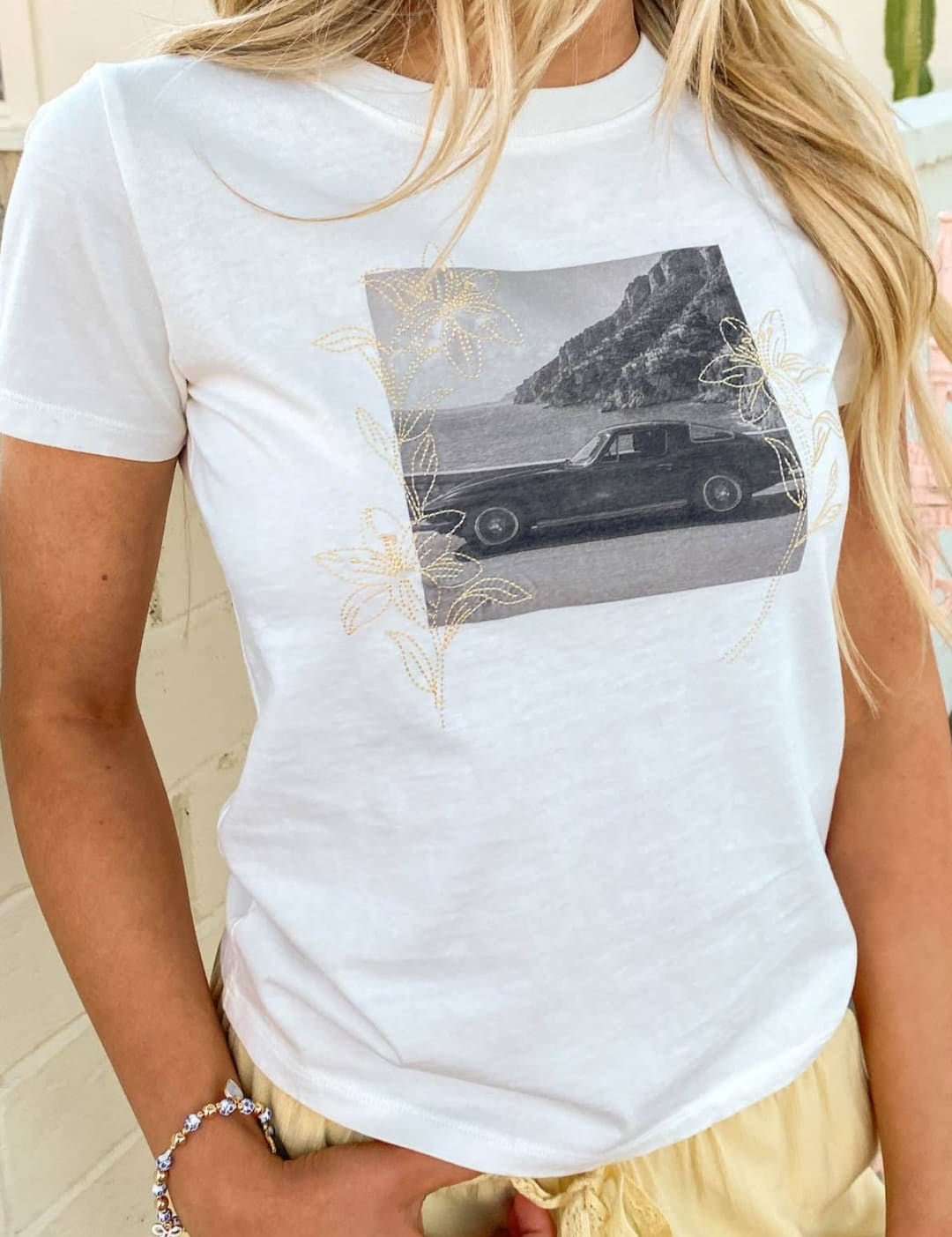 White short-sleeved t-shirt featuring a black-and-white graphic of a classic car on a coastal road, accented with delicate gold floral embroidery.