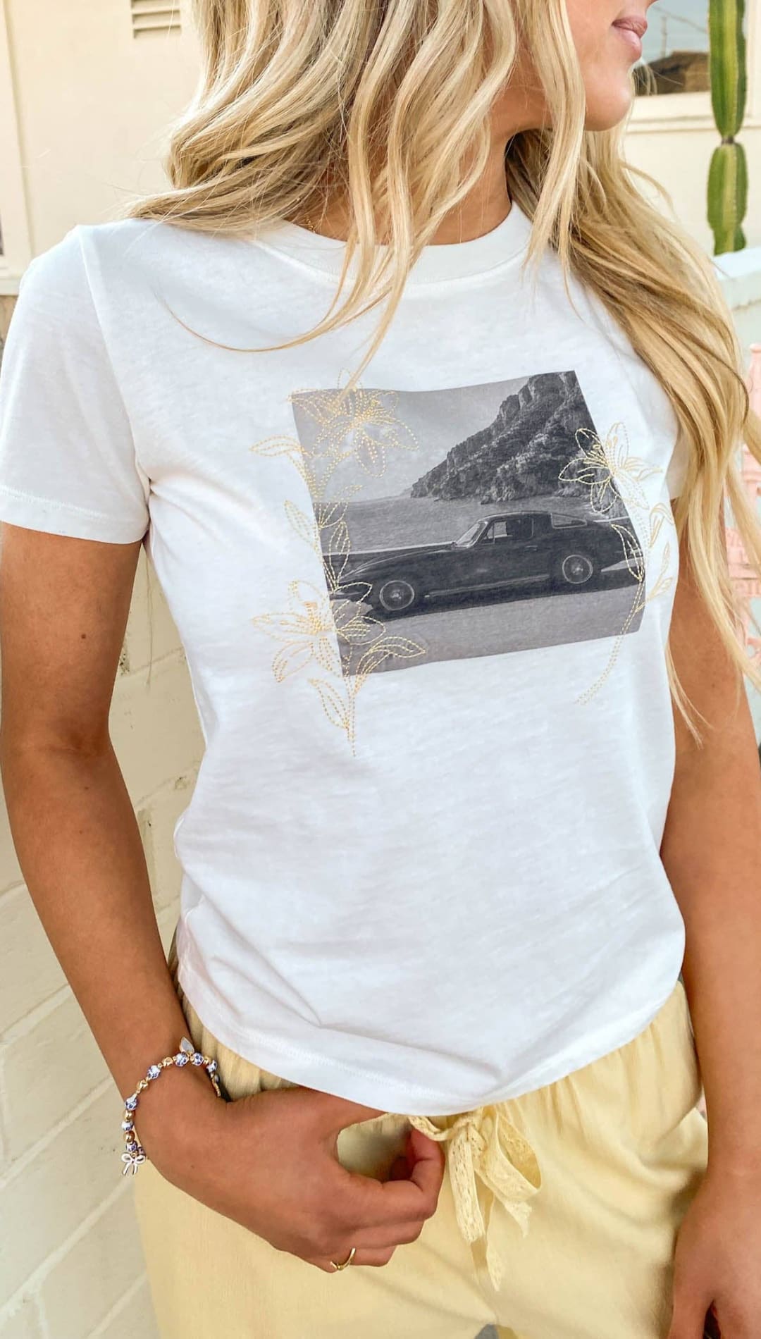 White short-sleeved t-shirt featuring a black-and-white graphic of a classic car on a coastal road, accented with delicate gold floral embroidery.