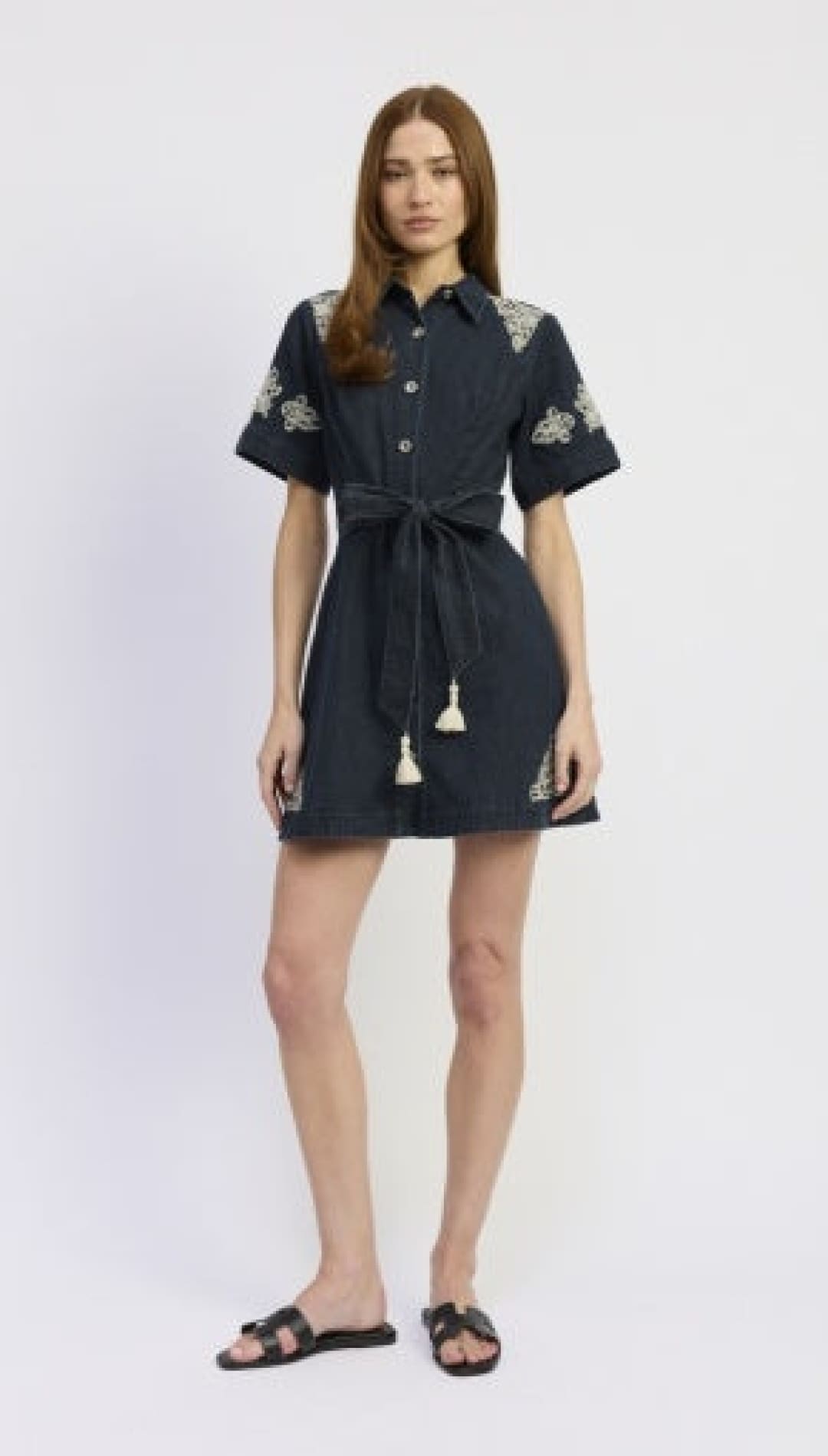 A navy blue denim dress with white floral embroidery on the sleeves and waistband, featuring a button-up front and a tied belt with tassel details.