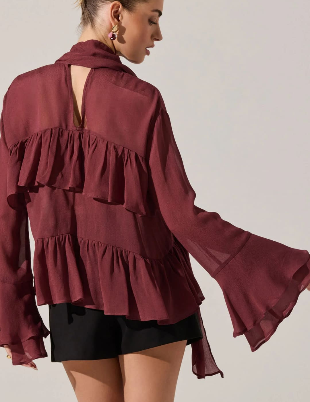 A sheer, tiered, ruffle-front blouse in a rich burgundy color, featuring a high collar with a keyhole back and long bell sleeves.