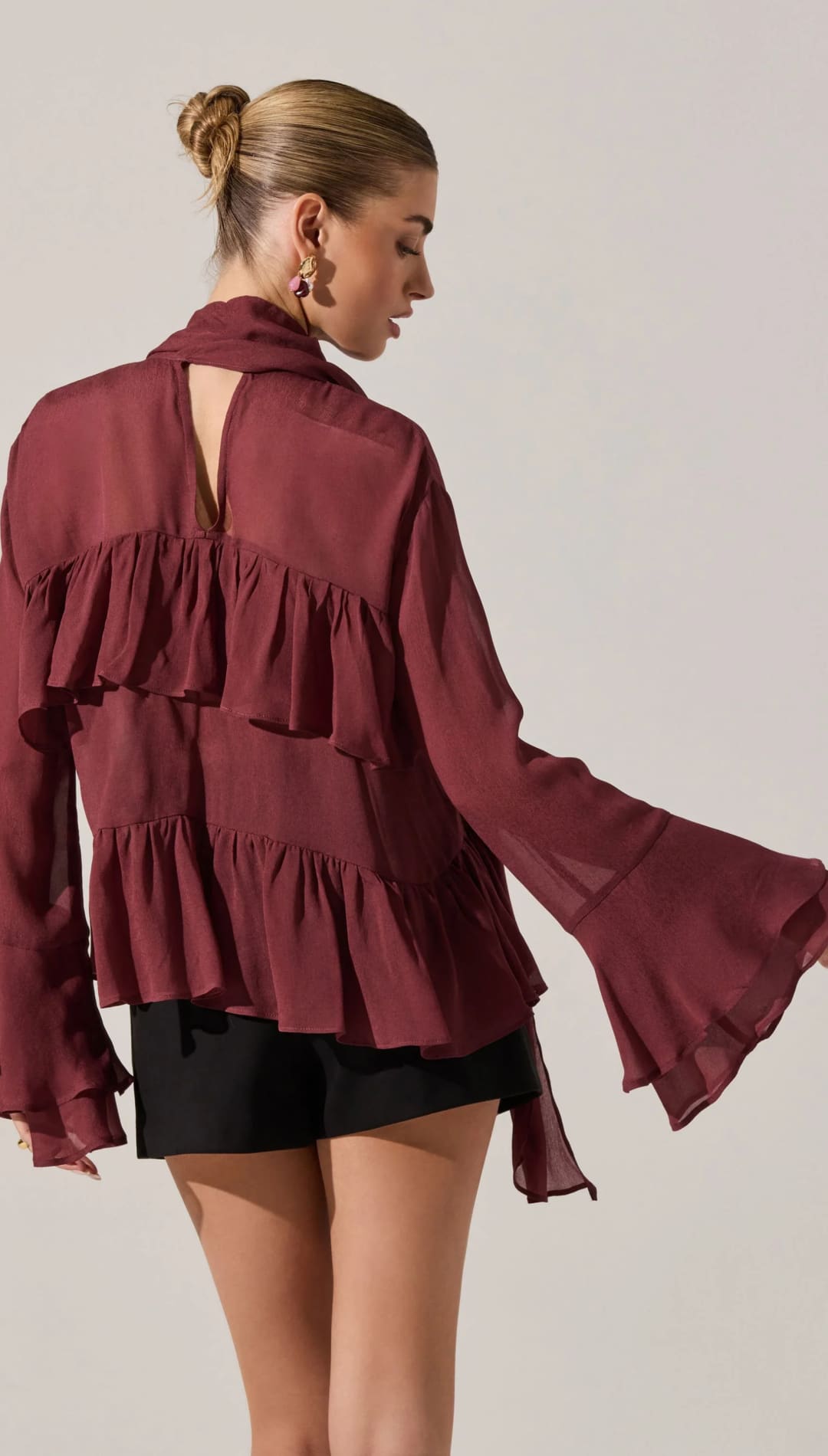 A sheer, tiered, ruffle-front blouse in a rich burgundy color, featuring a high collar with a keyhole back and long bell sleeves.
