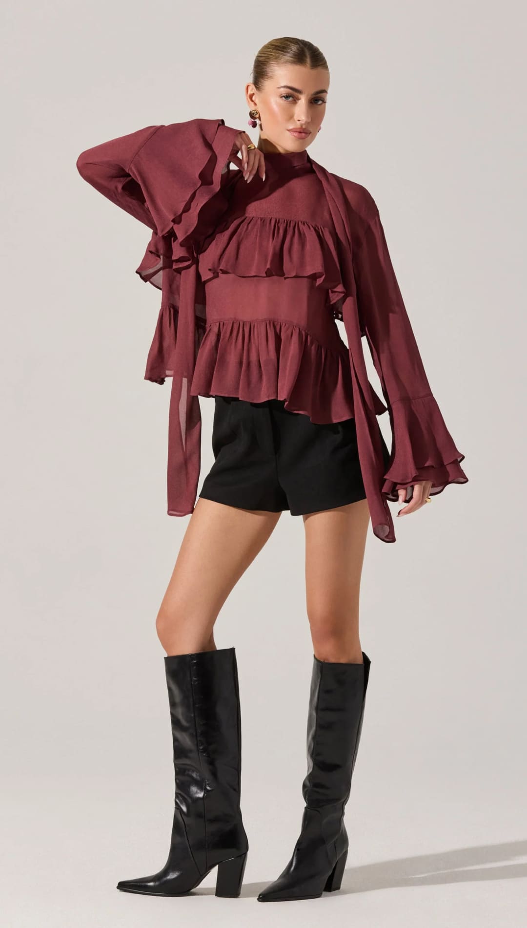 A long-sleeved maroon ruffled blouse with a high neck and tie detail, paired with black shorts and knee-high black boots.