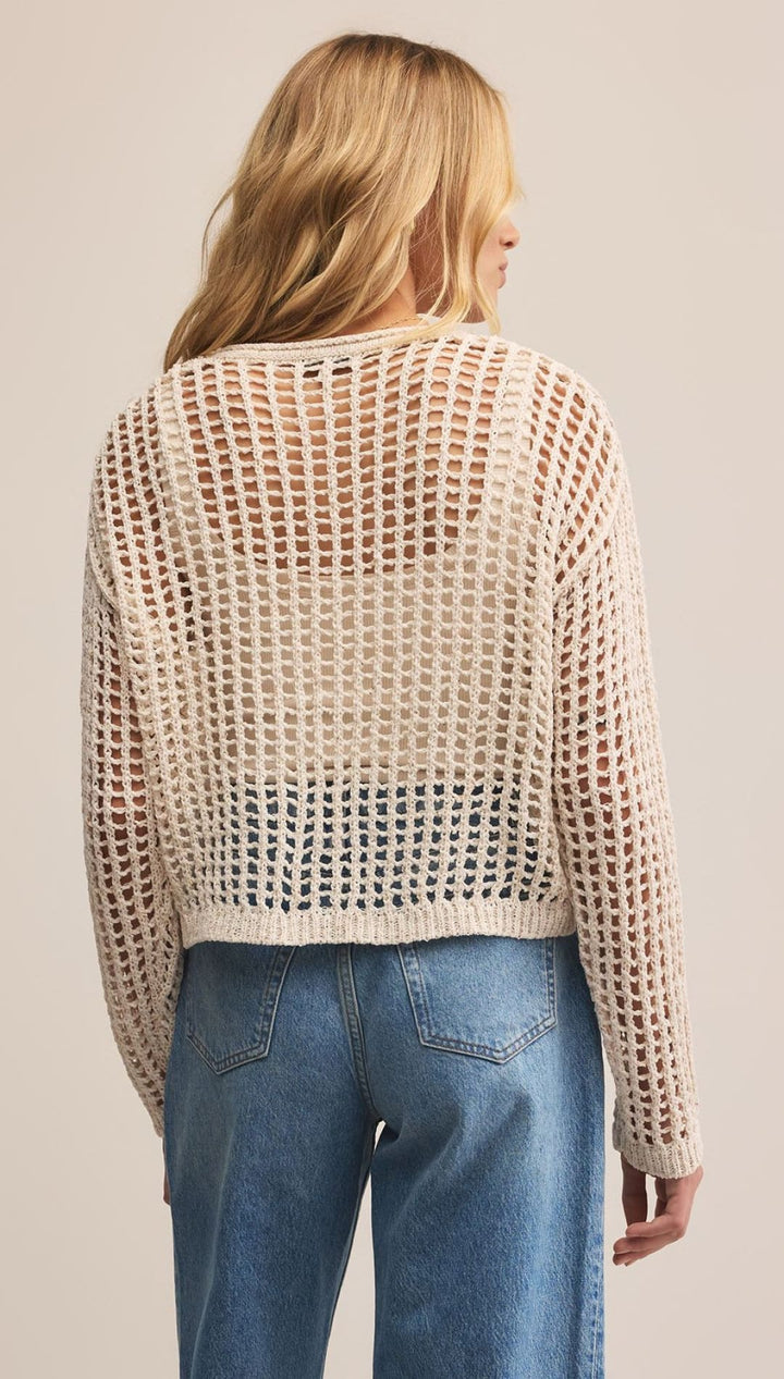 Off-white, open-knit, long-sleeved sweater worn over a white top and blue jeans.