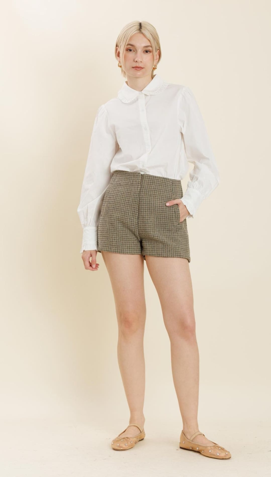 A woman models a white long-sleeved collared shirt with brown plaid shorts and nude ballet flats.
