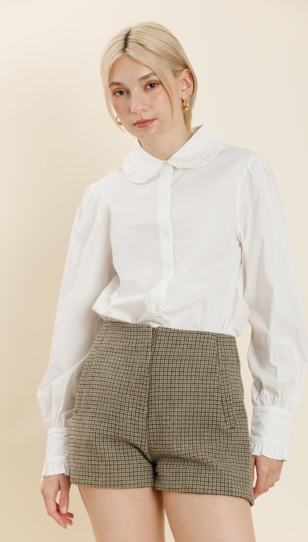 A woman’s white button-up shirt with a Peter Pan collar and puffed sleeves, paired with high-waisted, brown and black checkered shorts.