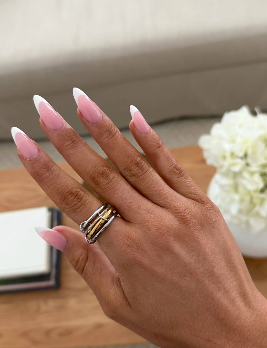 A hand with long, almond-shaped nails featuring a soft pink gradient french manicure and a silver ring with intertwined gold bands.