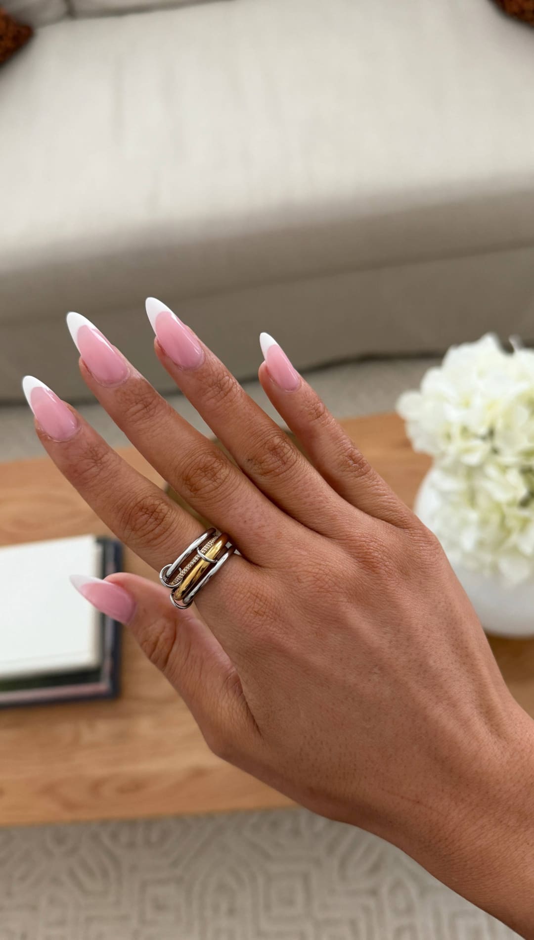 A hand with long, almond-shaped nails featuring a soft pink gradient french manicure and a silver ring with intertwined gold bands.