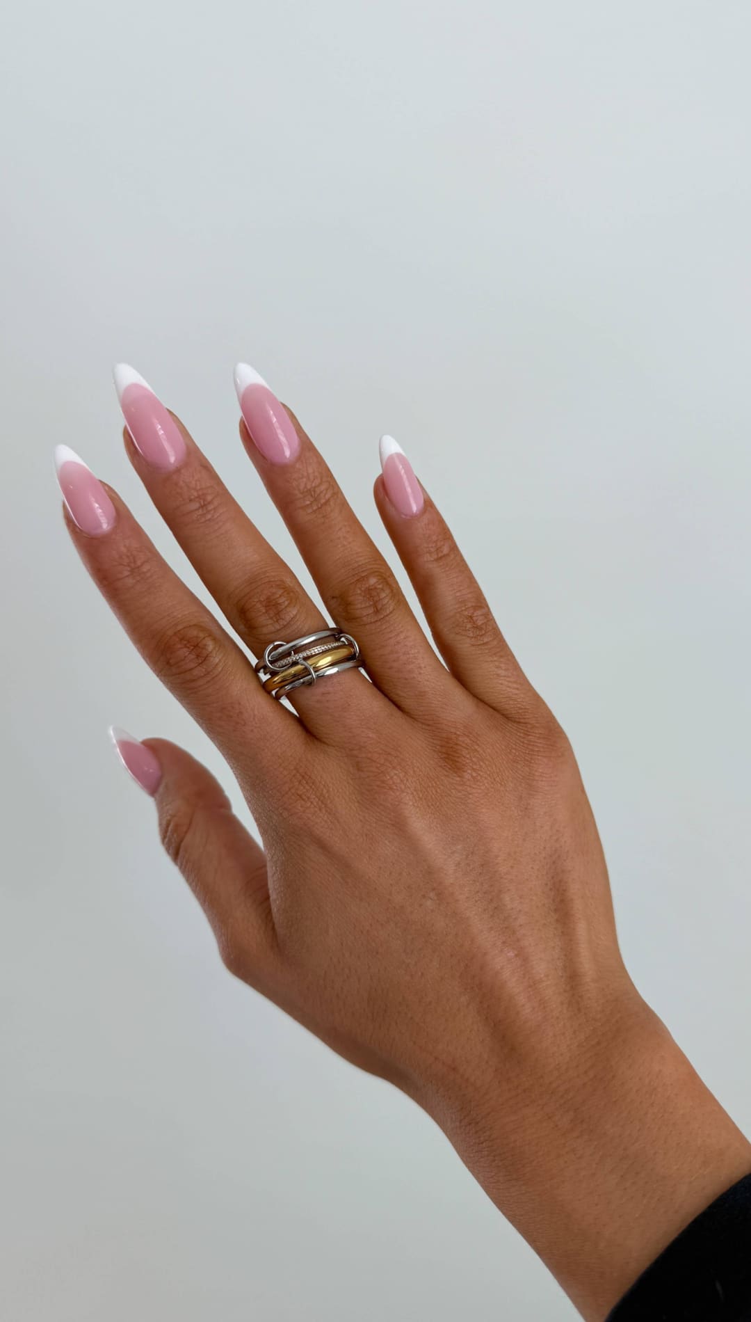 A hand with long, almond-shaped nails painted in a soft pink french manicure and wearing a multi-band ring made of interlocking silver-toned metal.