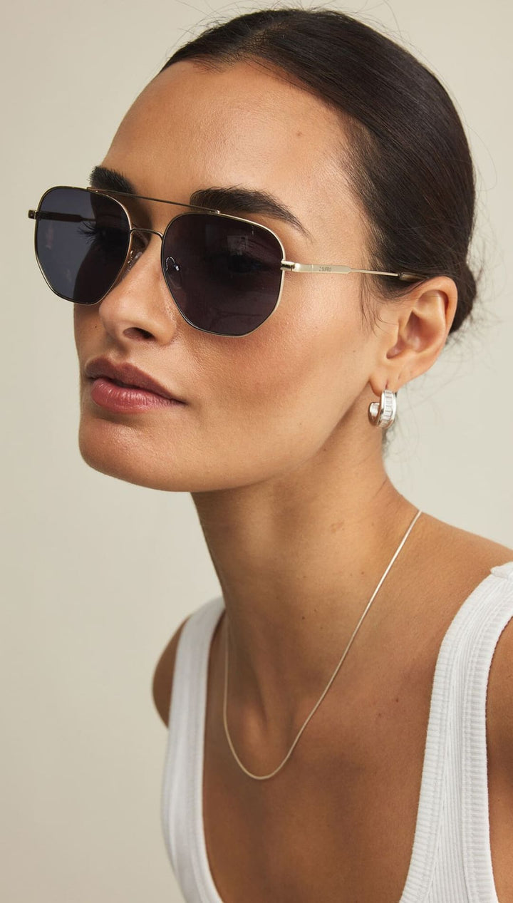 Woman wearing dark aviator-style sunglasses, a thin silver chain necklace, and small hoop earrings.