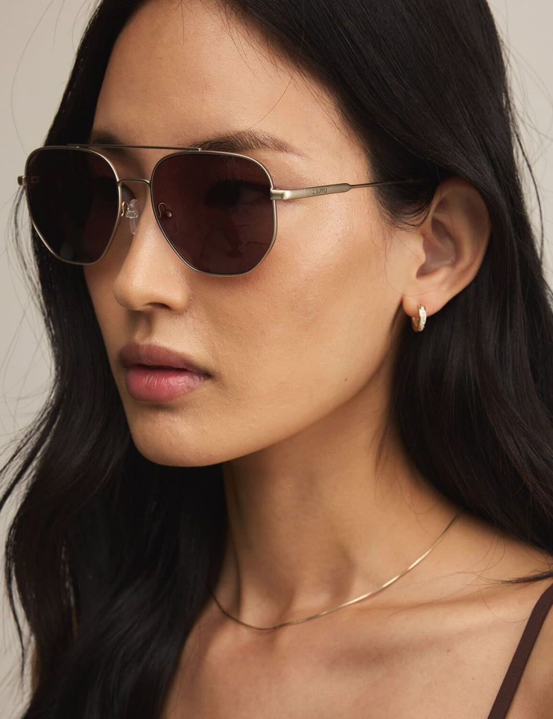 A woman with long dark hair wearing aviator sunglasses, a delicate gold chain necklace, and a brown top.
